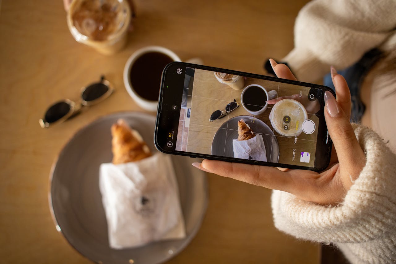 Woman photographing coffee and croissant with smartphone at a cafe.